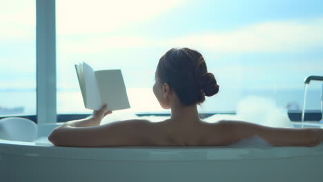 Back-of-sexy-woman-reading-the-book-in-a-bathtub-by-the-window,-lavish-lifestyle-concept,-full-frame