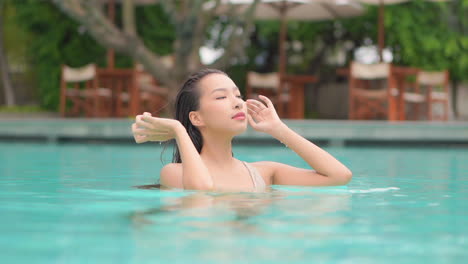 Young-sexy-asian-woman-caressing-her-wet-hair-in-swimming-pool,-looking-directly-to-camera,-slow-motion-full-frame