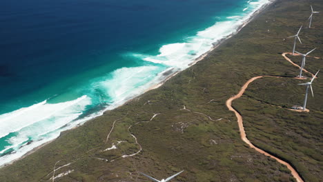 Aerial-view-of-waves-crashing-on-the-coastline-and-wind-power-turbines-rotating-in-the-wind,-in-Albany,-Australia---tracking,-drone-shot