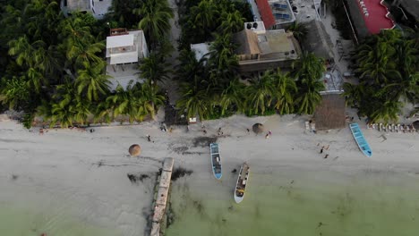 Bird’s-eye-view-ascending-shot,-Scenic-view-of-People-walking-on-the-shoreline-of-Isla-Holbox,-Mexico,-clear-beach-and-palm-tree-and-houses-in-the-background