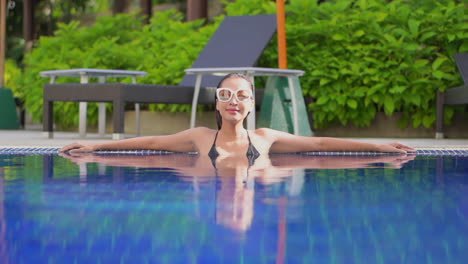 With-arms-stretched-out-along-the-edge-of-a-swimming-pool,-an-attractive-young-woman-turns-her-head-from-side-to-side
