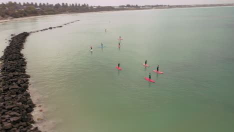Fast-aerial-circling-shot-showing-group-of-sup-paddlers-surfboarding-on-green-water-surface-in-Australia