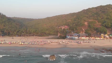Tourists-enjoying-the-ocean-in-Arambol-beach,-in-Goa,-India---Aerial-Fly-away-shot