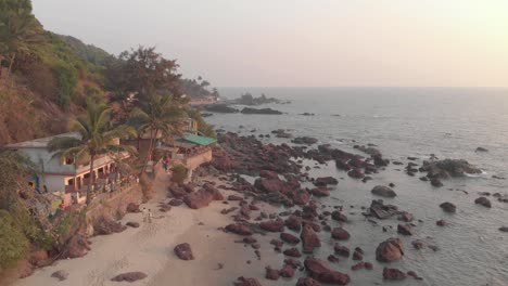Rocky-ocean-coast-and-old-buildings-in-Arambol-Beach-at-sunset-in-Goa,-India---Aerial-Fly-over-shot