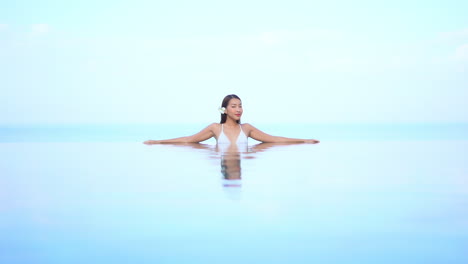 Mysterious-sexy-Asian-woman-inside-the-infinity-pool-slowly-turning-her-sight-towards-camera-on-the-simplistic-sky-background,-template