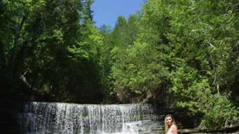 Camera-tilts-down-to-reveal-a-young-woman-in-a-bikini-standing-in-a-river,-waterfall-in-background