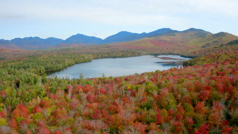 Aerial-Pull-Back-Shot-of-a-Lake-Surrounded-by-Beautiful-Fall-Foliage-with-a-Mountain-Ridge-in-the-Background-on-a-Sunny-Day