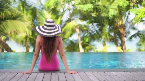 Back-view-of-young-woman-with-large-hat-and-pink-swimsuit-sitting-at-pool-edge