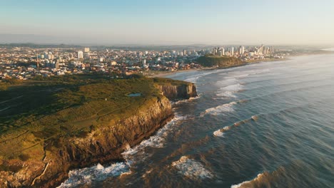 Aerial-view-of-high-rocky-cliffs-by-atlantic-ocean-and-city-skyline-on-the-back,-Torres-City,-Rio-Grande-do-Sul,-Brazil