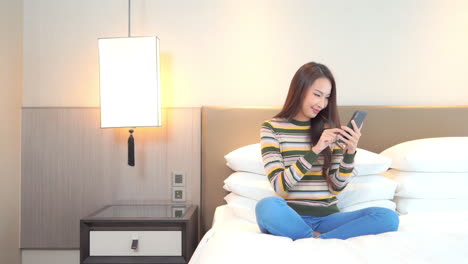 Woman-chats-on-smartphone-while-sitting-with-legs-crossed-on-bed