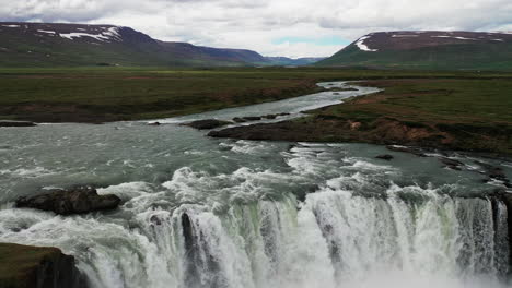 Man-Standing-In-Front-Of-Godafoss-Waterfall-With-Rushing-Waters-Flowing-From-The-River-In-North-Iceland---Drone-Shot