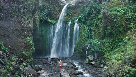 Woman-traveler-in-bikini-stands-at-magical-Tiu-Kelep-waterfall-in-Lombok,-aerial