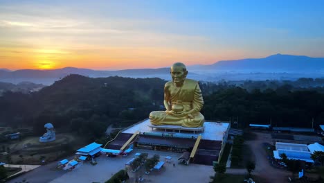 4k-Wide-Aerial-shot-of-a-Big-Luang-Por-Tuad-monk-statue-surrounded-by-mountains-of-Khao-Yai-at-dusk-in-Thailand