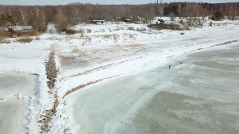Drone-View-of-Snow-Covered-Beach,-Closing-in-on-People-Walking-on-Shore