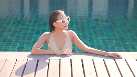 A-young-woman-fresh-from-a-dip-in-the-pool-stands-alone-in-the-pool-resting-her-arm-on-the-wooden-deck-surrounding-the-pool