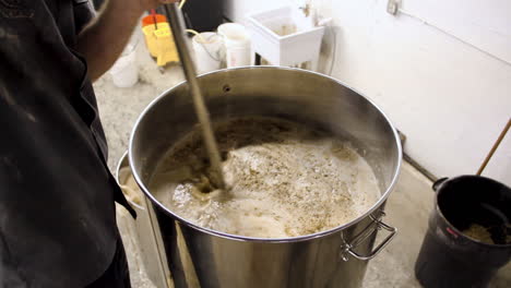 Worker-in-beer-factory-pouring-fresh-produced-beer-with-wheat-and-liquid-in-large-pot