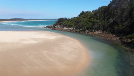 Wide-rotating-drone-shot-of-the-South-Pacific-Ocean,-Korogoro-Creek-and-inlet-with-wind-blowing-sand-across-a-sand-bar-at-Hat-Head-New-South-Wales,-Australia