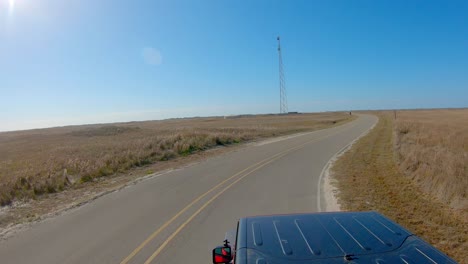 Vehicle-top-POV-while-driving-thru-a-curve-on-paved-road-thru-grasslands-and-towards-cell-tower