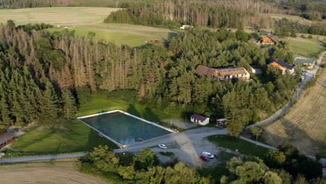 Aerial-Shot-Of-Luxury-Outdoors-Swimming-Pool-Surrounded-By-Farm-Land-And-Forest-At-Sunrise