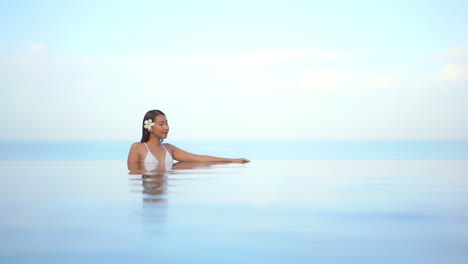 Young-beautiful-woman-wearin-a-swimsuit-leaning-of-the-edge-of-an-infinity-pool-with-blurred-ocean-and-blue-sky-in-the-background