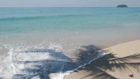 Kleine-Wellen-Des-Tropischen-Ozeans-Umspülen-Das-Weiße-Sandstrand-Mit-Palmenschatten-Und-Insel-Im-Hintergrund