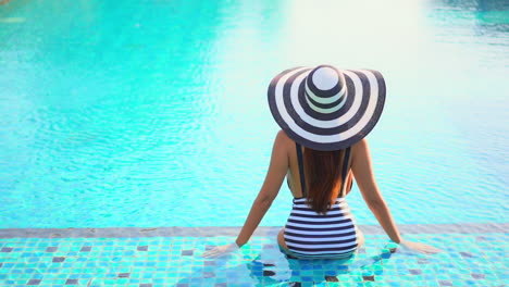 Rear-Shot-of-Fashionable-Woman-Sitting-at-Shallow-End-of-Swimming-Pool