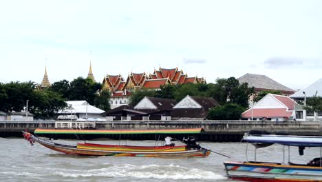 View-of-beautiful-temple-on-the-bank-of-a-busy-river-with-tourist-and-traditional-boats-passing-by