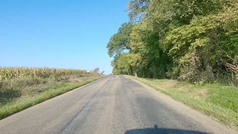 POV-driving-on-rural-county-road-with-no-center-line-past-maturing-fields,-farmyard,-trees-and-a-cemetery-in-rural-Iowa-on-a-sunny-early-autumn-day
