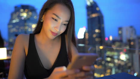 Attractive-Trendy-Asian-Woman-Using-Smartphone-on-Rooftop-Terrace,-Nighttime-City-Background-CLOSE-UP