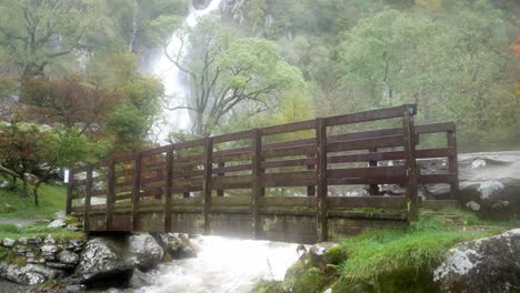 Idyllic-wooden-bridge-over-misty-wet-valley-waterfall-cascading-into-powerful-flowing-river-close-up