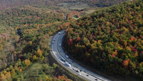 Traffic-rounds-corner-through-mountains-covered-in-autumn-fall-foliage,-colorful-leaves,-trucks-and-cars-on-interstate-turnpike-highway-road