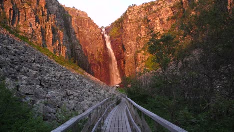 wide-shot-from-afar-of-majestic-Njupeskärs,-Sweden's-highest-waterfall-lit-by-midsummer-morning-golden-sun,-guided-by-wooden-passageway-in-Fulufjällets-Nationalpark