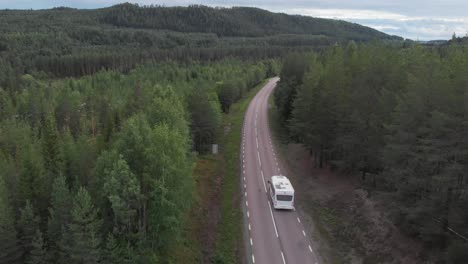 Aerial-wide-shot-tracking-white-camper-van-adventuring-in-green,-dense,-coniferous-trees,-riding-down-endless-lonely-mountain-road