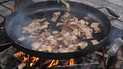 Close-up-high-angle-tilt-down-shot-of-kebab-beef-and-lamb-meat-being-stir-fried-on-muurikka-griddle-pan,-over-a-hot-wood-fire-in-a-camping-environment