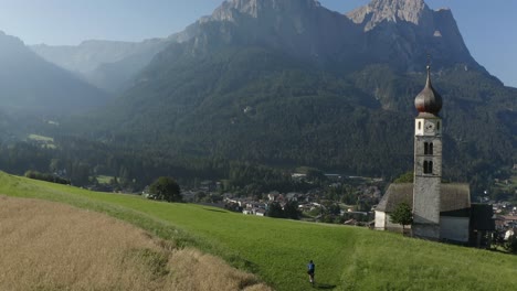 Aerial-following-a-cyclist-going-through-a-green-field-in-the-Italian-Alps-with-a-Church-and-Mountain-background,-Kastelruth,-St