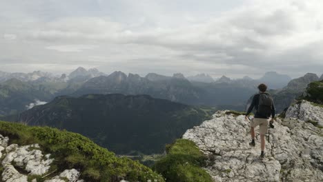 Epic-man-adventurer-walks-up-to-a-cliff-ridge-to-have-a-beautiful-view-vista-in-mountain-backdrop,-Alleghe,-Refugio-Tissi,-Italy