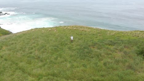 Aerial-orbit-around-a-man-walking-in-green-grass-on-the-edge-of-a-large-cliff-with-the-big-southern-ocean-in-view