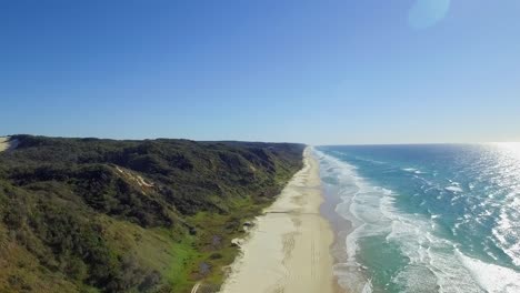 Stunning-aerial-view-along-the-broad-beach-of-Fraser-Island,-K�gari,-with-the-wooded-slopes-of-its-huge-dunes-and-the-sun-sparkling-off-the-ocean-on-a-hot-summer�s-day