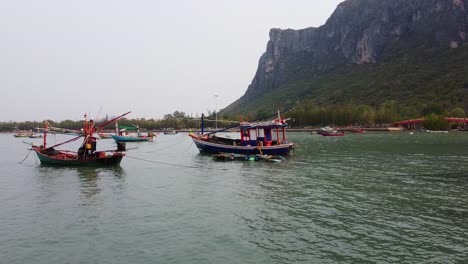 Drone-travels-forward-to-Asian-colorful-fishing-boats-at-Khao-Ta-Mong-Lai-Bay-with-rocky-Mountain-on-the-background