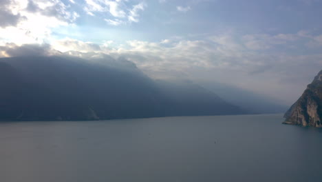 Dolly-out-aerial-shot-of-Lake-Garda-with-the-morning-sun-breaking-through-the-clouds-and-spilling-light-over-the-majestic-mountains