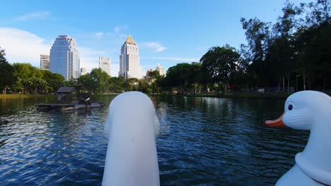 POV-footage:-Heads-of-a-Swan-paddle-boat-on-the-lake-with-Skyscrapers-on-the-background-in-Lumphini-Park,-Bangkok,-Thailand