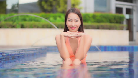 Sexy-Asiatische-Frau,-Die-Langsam-Ihren-Blick-In-Richtung-Der-Kamera-Dreht-Und-Lächelt,-Während-Sie-Im-Flachen-Schwimmbadwasser-Im-Hotelresort-Sitzt,-Nahaufnahme,-Zeitlupe