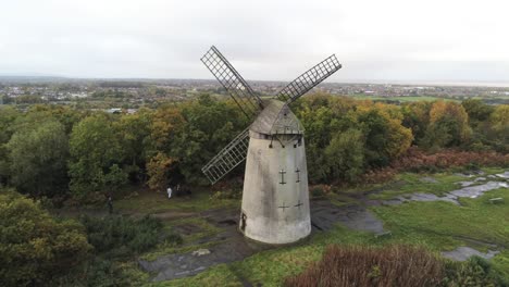 Traditional-wooden-stone-flour-mill-windmill-preserved-in-Autumn-woodland-aerial-view-countryside-rising-forwards-tilt-down-birdseye