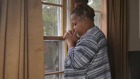 Black-adult-woman-standing-in-front-of-window-on-a-rainy-day-praying-to-God-for-better-days-in-this-world