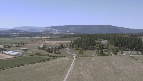 aerial-fly-over-industrial-farm-land-bare-mountains-within-a-valley-with-curvy-roads-tall-pine-trees,-dairy-wheat-cattle-barns-on-the-countryside-in-British-Columbia-off-the-freeway-to-Alberta-CA-1-5