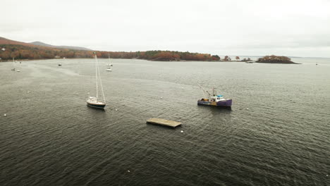 Lobster-boat-and-sailboat-anchored-in-bay,-foggy-fall-day,-drone-shot
