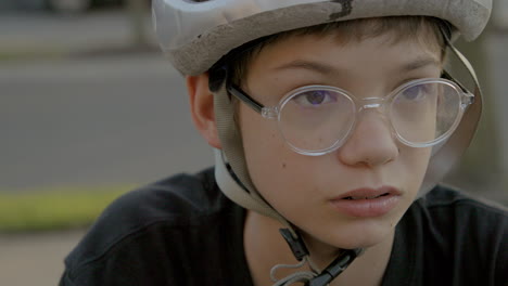 Cute-teen-kid-with-glasses-and-a-bike-helmet-sitting-outside-looks-down-and-then-looks-up-and-off-camera