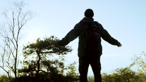 Silhouette-of-man-with-backpack-in-wilderness-pulling-up-arms-in-front-of-sunlight-in-nature