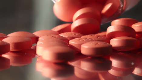 Many-small-pink-red-prescription-drug-pills-tablets-falling-from-bottle-onto-dark-mirrored-surface-close-up
