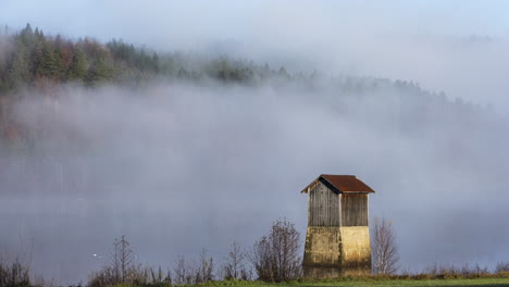 Small-wooden-structure-at-edge-of-fog-covered-lake,-abandoned-timber-cable-car